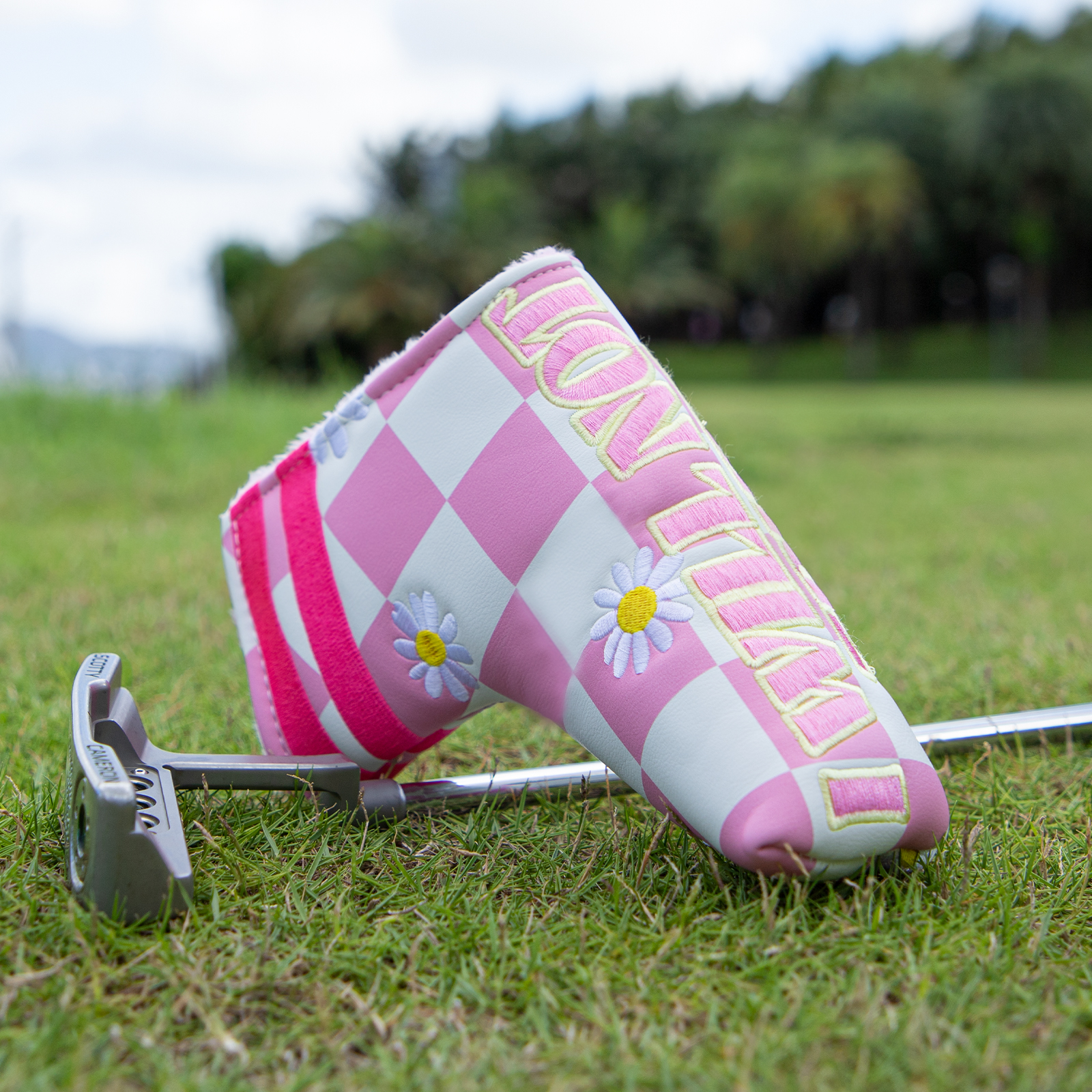 Two pink checkerboard covers with daisy graphics are shown on grass beside golf clubs and club heads.
