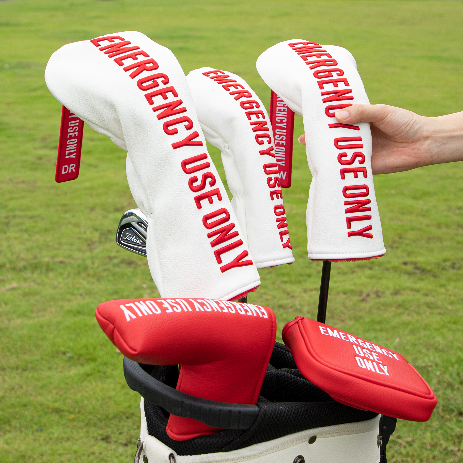 A hand holds three white covers above a golf bag on grass; red “EMERGENCY USE ONLY” lettering is visible.